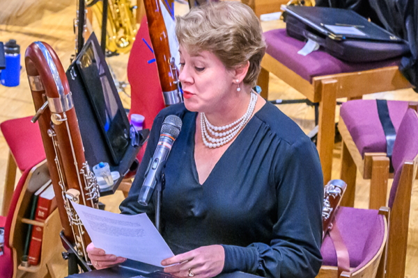 Woman with short blonde hair, wearhing a dark blue long-sleeved dress and a white, four-strand pearl necklance. She is holding a sheet of paper and is speakiong into a silver microphone.