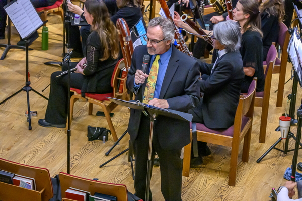 Man with glasses and curly graying hair is speaking into a microphone held in his right hand. He is wearing a dark blue suit, brighter blue collared shirt, and a yellow patterned tie.