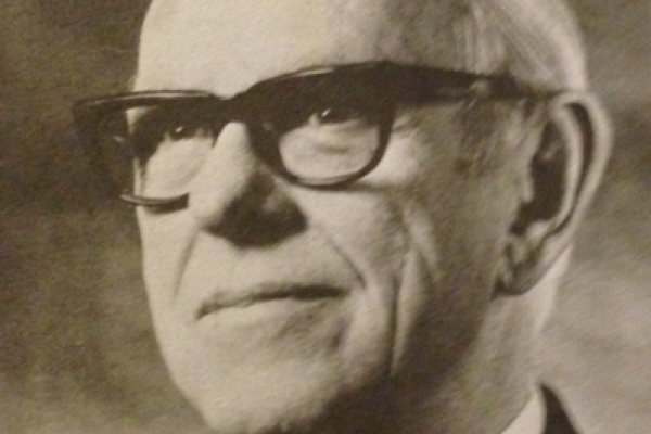 sepia-tone image of balding man in dark suit and striped tie, wearing dark-framed glases